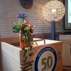 Valtuustosalissa puhujapönttö, jonka päällä kukka-asetelma. Edessä pyöreä merkki, jossa lukee Säkylän kansalaisopisto 50 vuotta.